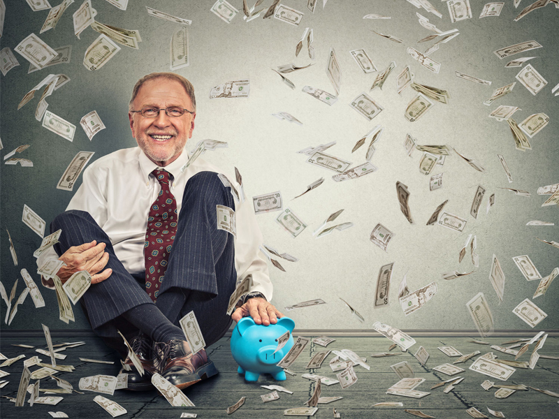 Excited happy senior business man sitting on a floor with piggy bank under a money rain isolated on gray wall background.