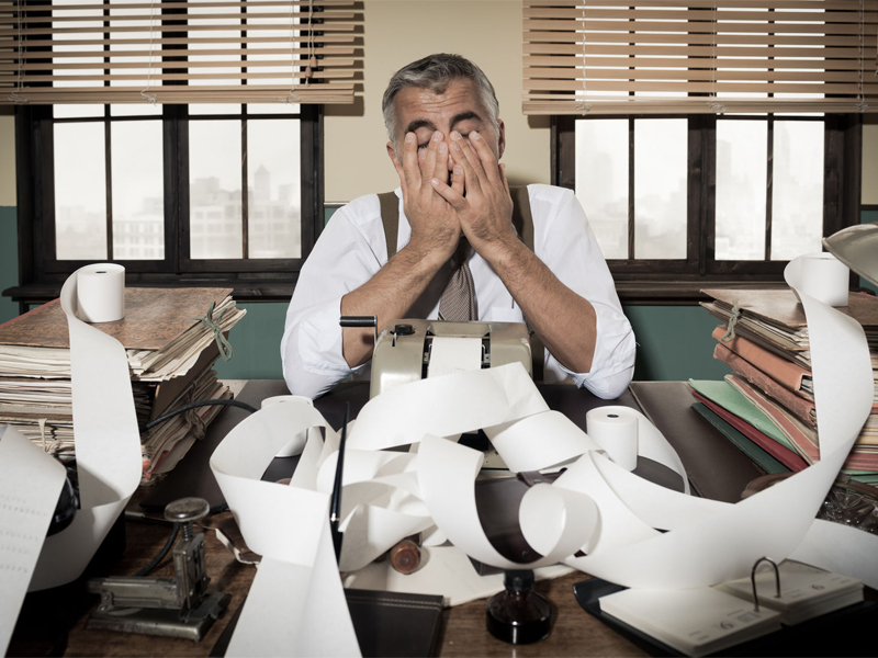 Desperate accountant head in hands surrounded by bills on paper tape, 1950s style office