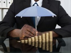 businessman protecting stack of coins with umbrella at desk