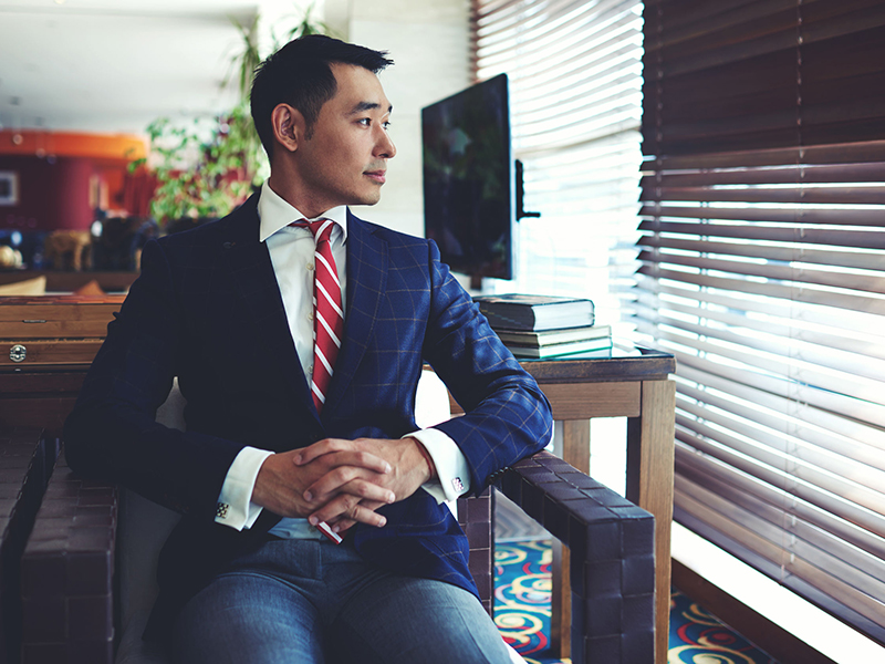 Portrait of young confident asian businessman with serious face sitting in modern office interior near big window, intelligent men entrepreneur in elegant suit thinking about something before meeting dream big business planning advisor