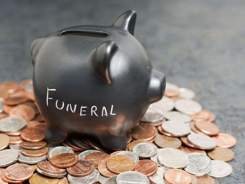 A coin piggy-bank with "Funeral" written on it sitting on a pile of coin.