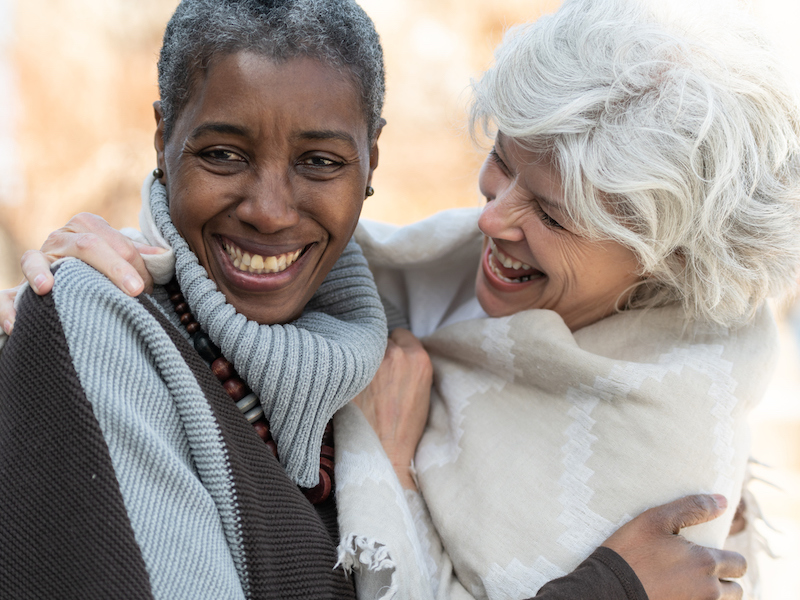 Two senior friends laugh affectionately together. The women are standing outside on a sunny but cool day. They are dressed in casual sweaters. One woman is of African descent and the other is Caucasian.