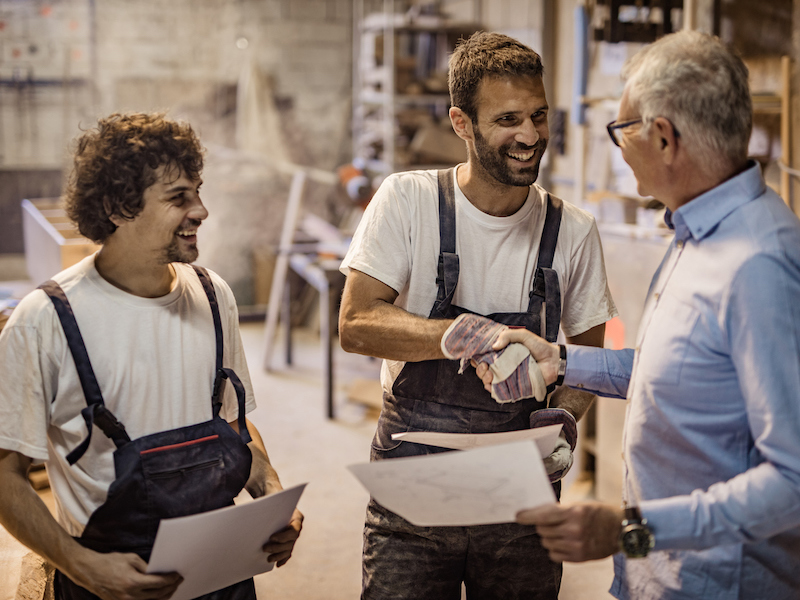 Two happy carpenters shaking hands with senior customer after successful agreement in a workshop.