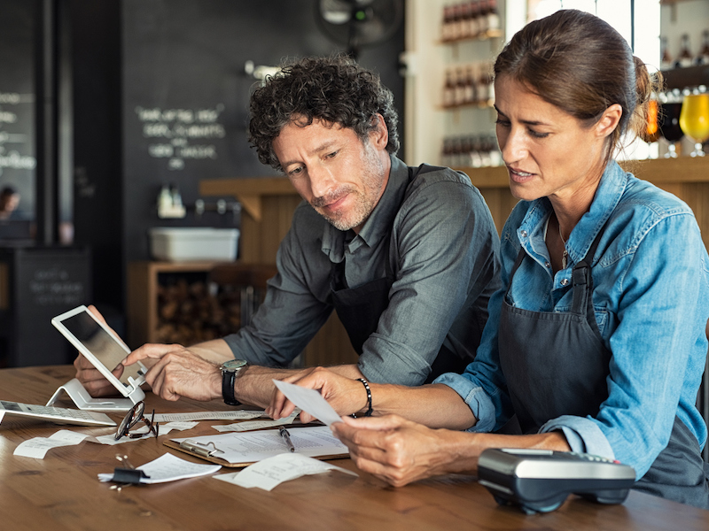 Man and woman sitting in cafeteria discussing finance for the month. Stressed couple looking at bills sitting in restaurant wearing uniform apron. Cafe staff sitting together looking at expenses and bills. (Man and woman sitting in cafeteria discussi