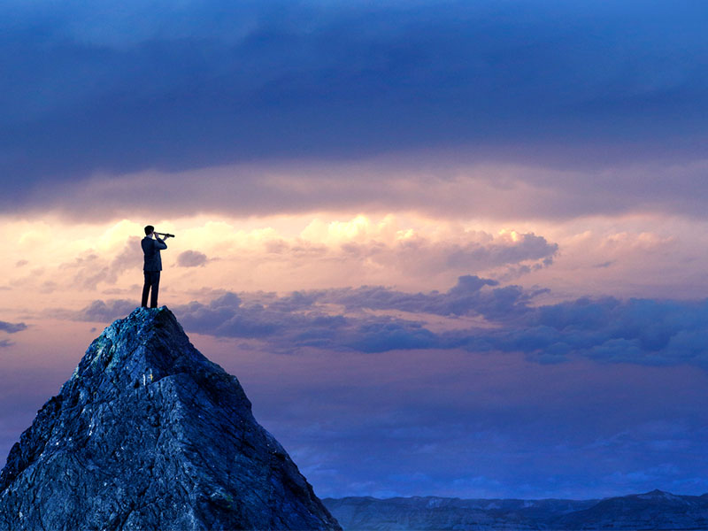 A silhouetted businessman stands on top of a mountain peak and looks into the distance with a spyglass in front of a dramatic sunset in the distance