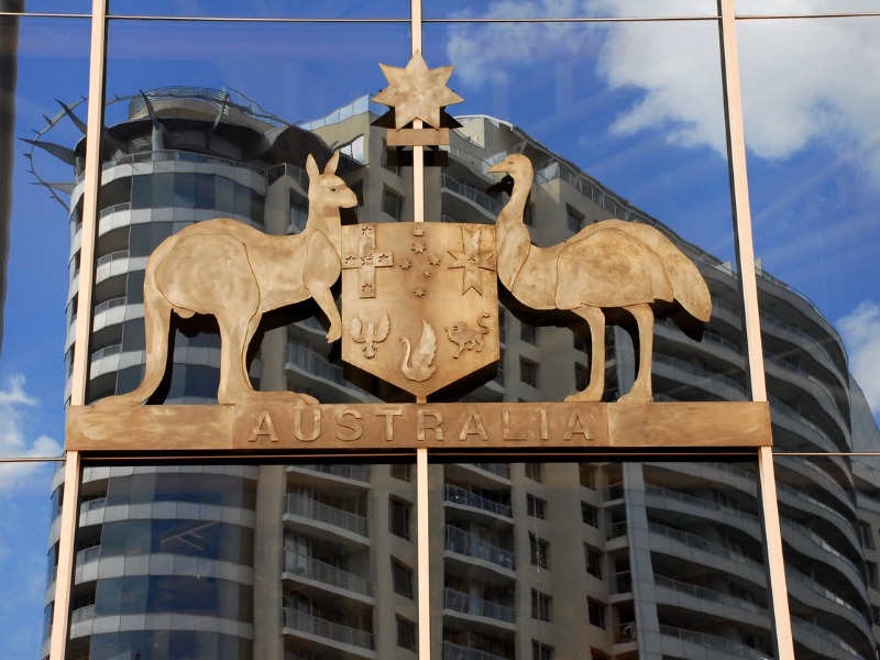 The Australian Coat of Arms in metal hangs on the glass wall of a building that reflects another high-rise across the street.