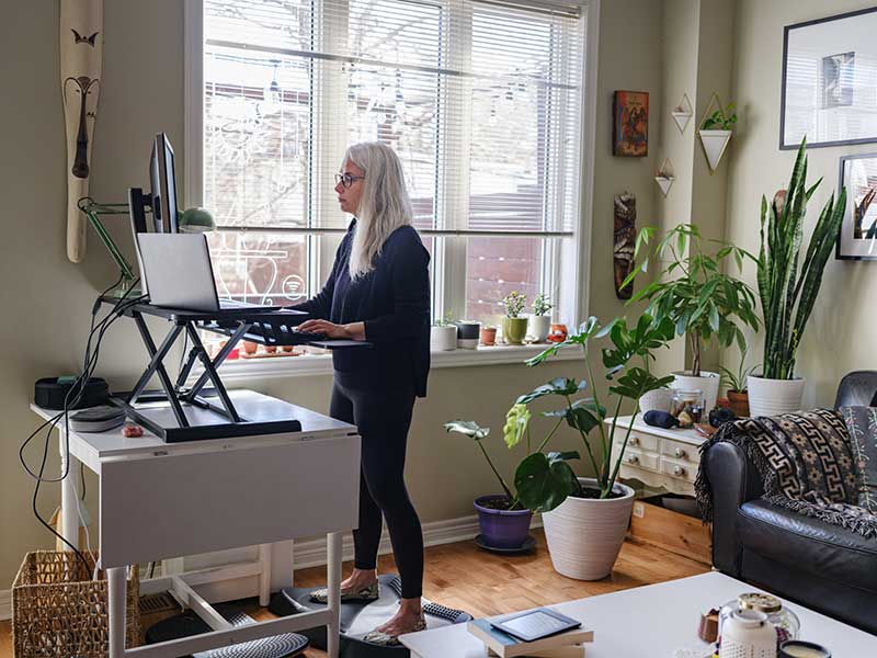 Life in time of COVID-19: Mature woman working on computer from home at the stand up desk. She has long grey hair and dressed in casual black outfit. Interior of living room set up with home office station, next to window.