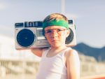 A young boy dressed in headband and retro workout attire is having a great time listening to music on his boombox.