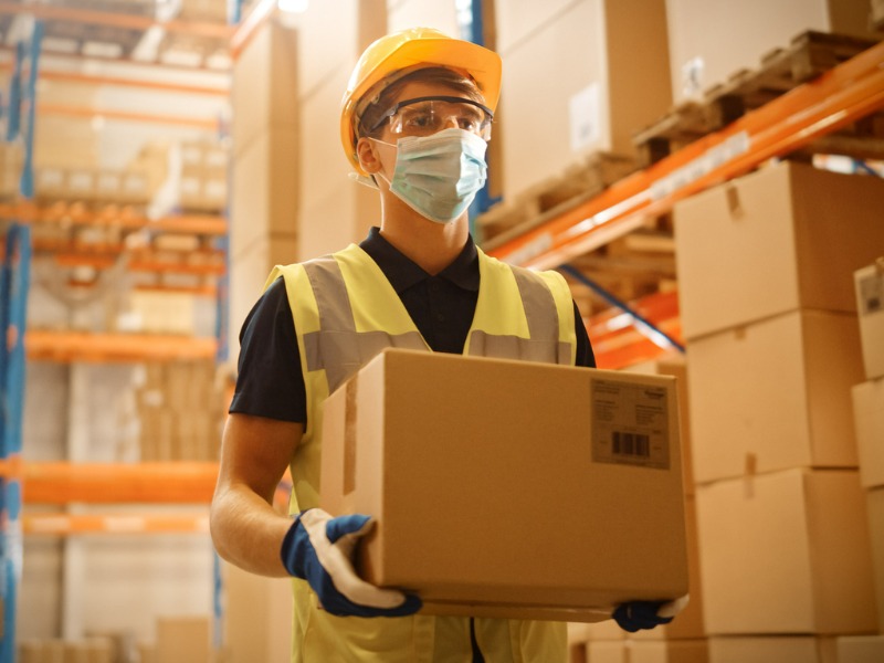 Worker Wearing Medical Face Mask and Hard Hat Carries Cardboard Box Walks Through Retail Warehouse full of Shelves with Goods.