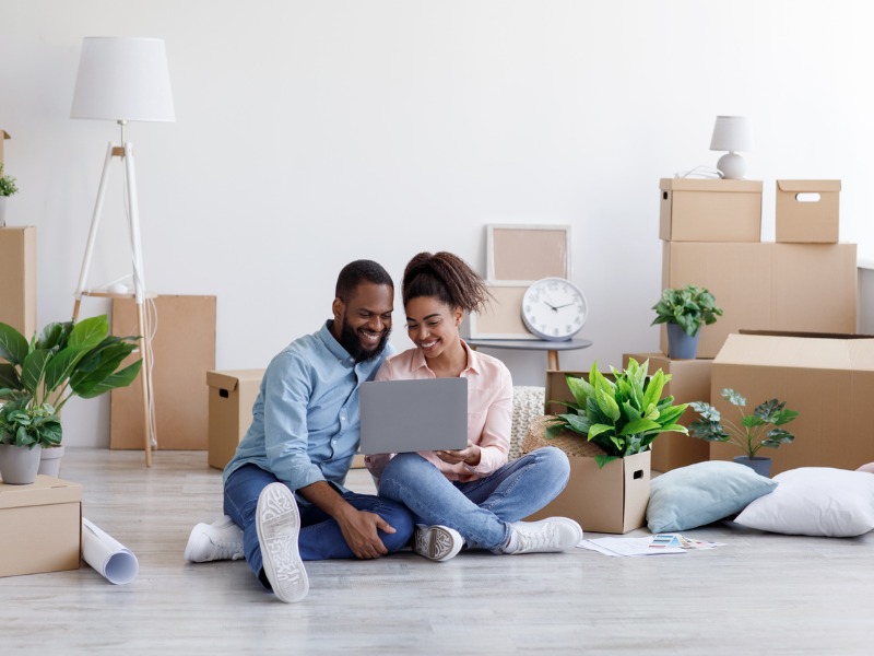 A young couple sitting on the floor among cardboard boxes with things and plants in the new home and looking at laptop.
