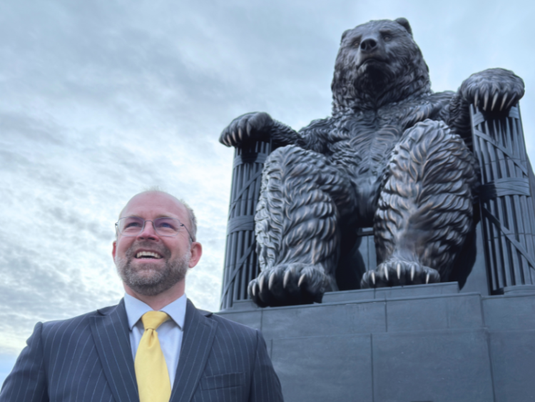Man in a suit smiling in front of a large seated bear statue against a cloudy sky.